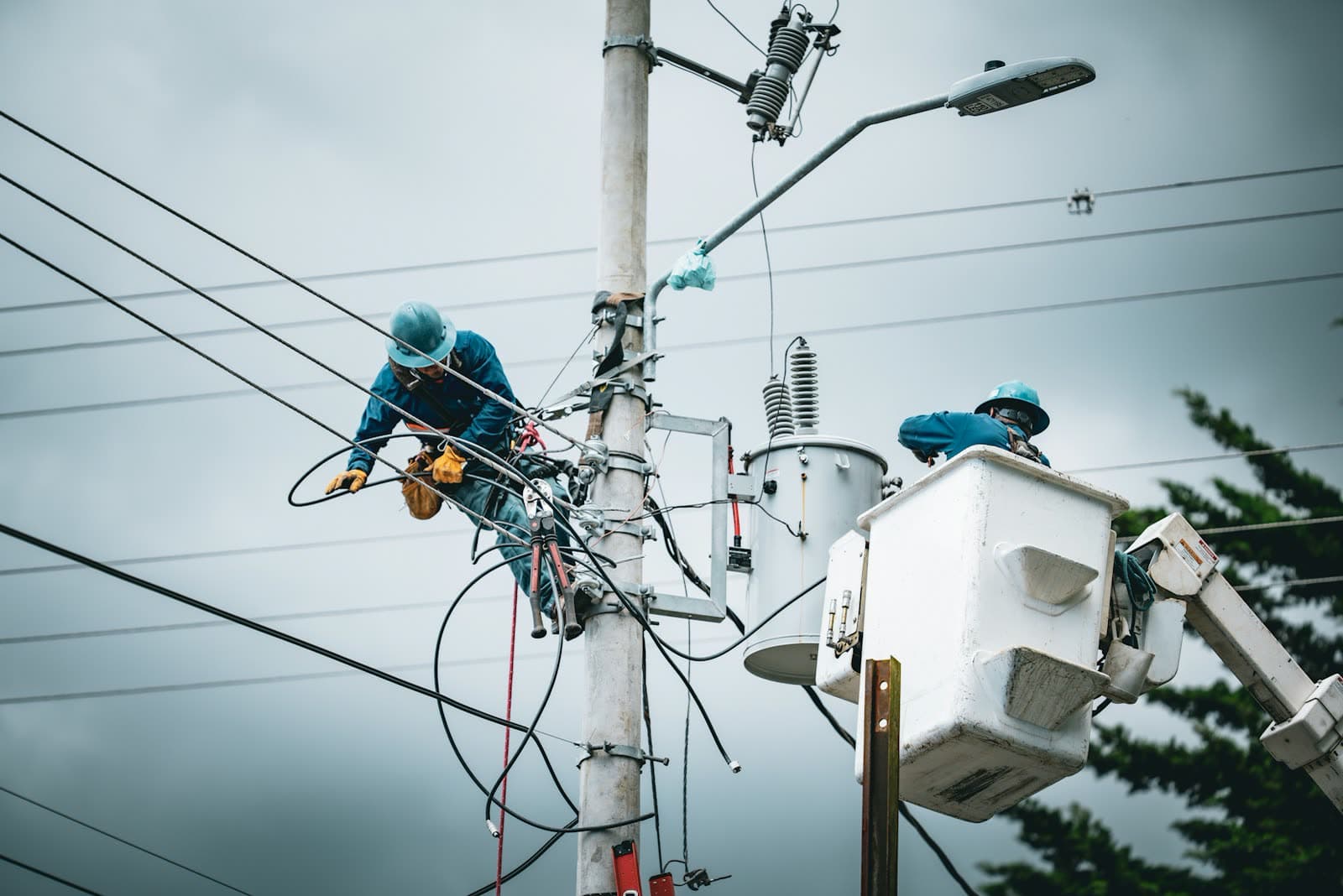 Two line workers repairing overhead power cables from a pole and a bucket lift under an overcast sky