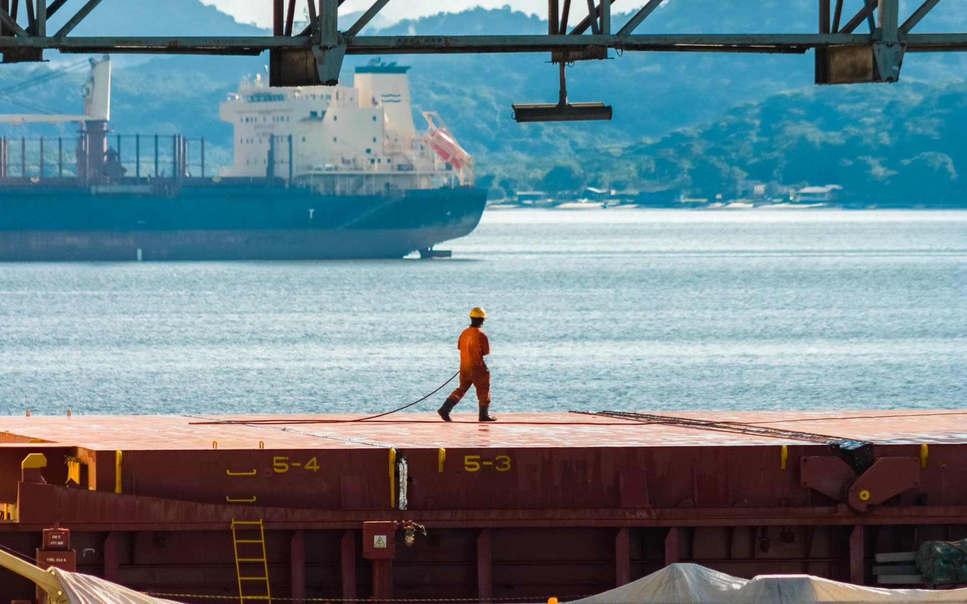 A dock worker in an orange suit crossing a barge deck, with a cargo ship and hills beyond